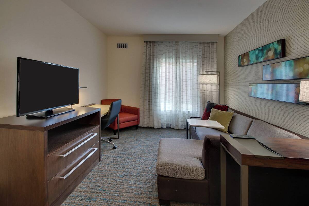 A hotel room with a flat-screen TV on a wooden stand, a red armchair, a sofa, a small table, and a window with sheer curtains.