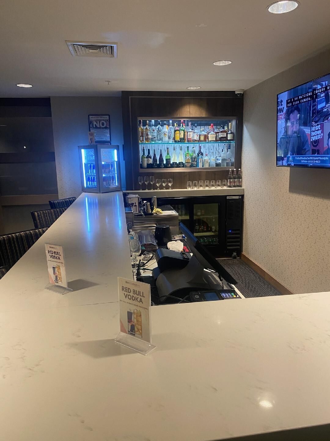 A bar area with a long counter, a television screen, and a variety of bottles on shelves.