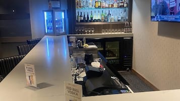 A bar area with a long counter, a television screen, and a variety of bottles on shelves.
