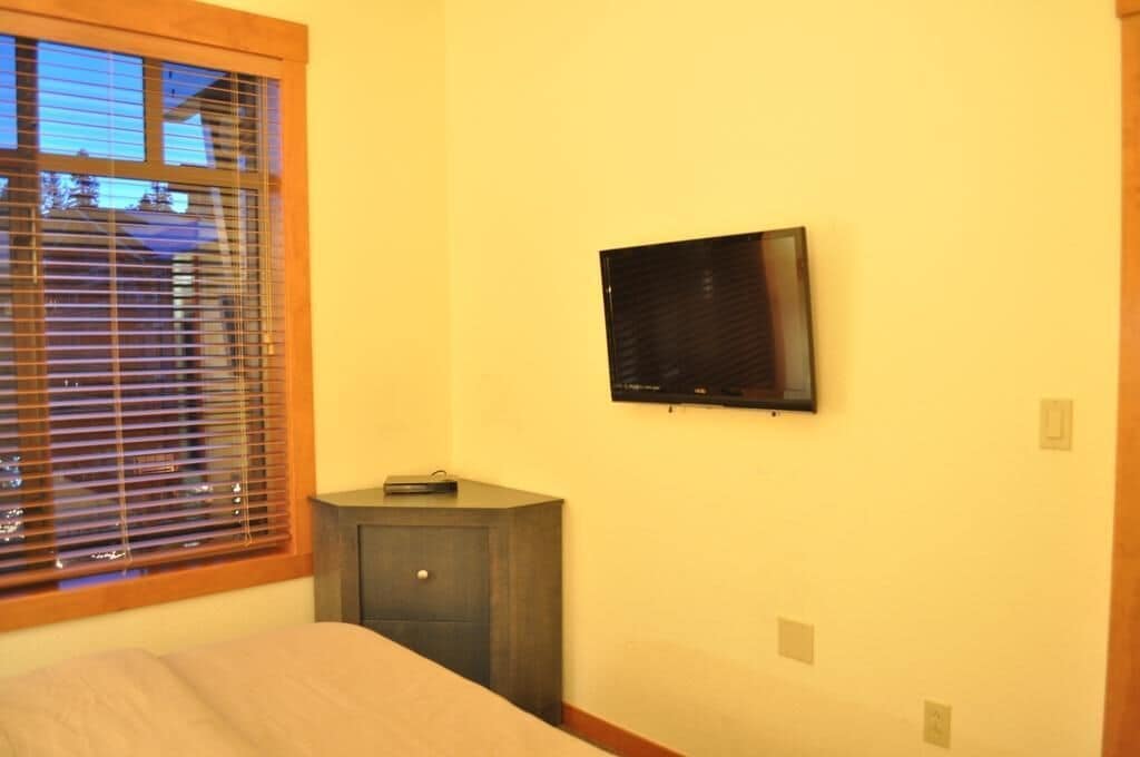 A bedroom with a bed, a small bedside table, a TV mounted on the wall, and a window with blinds.