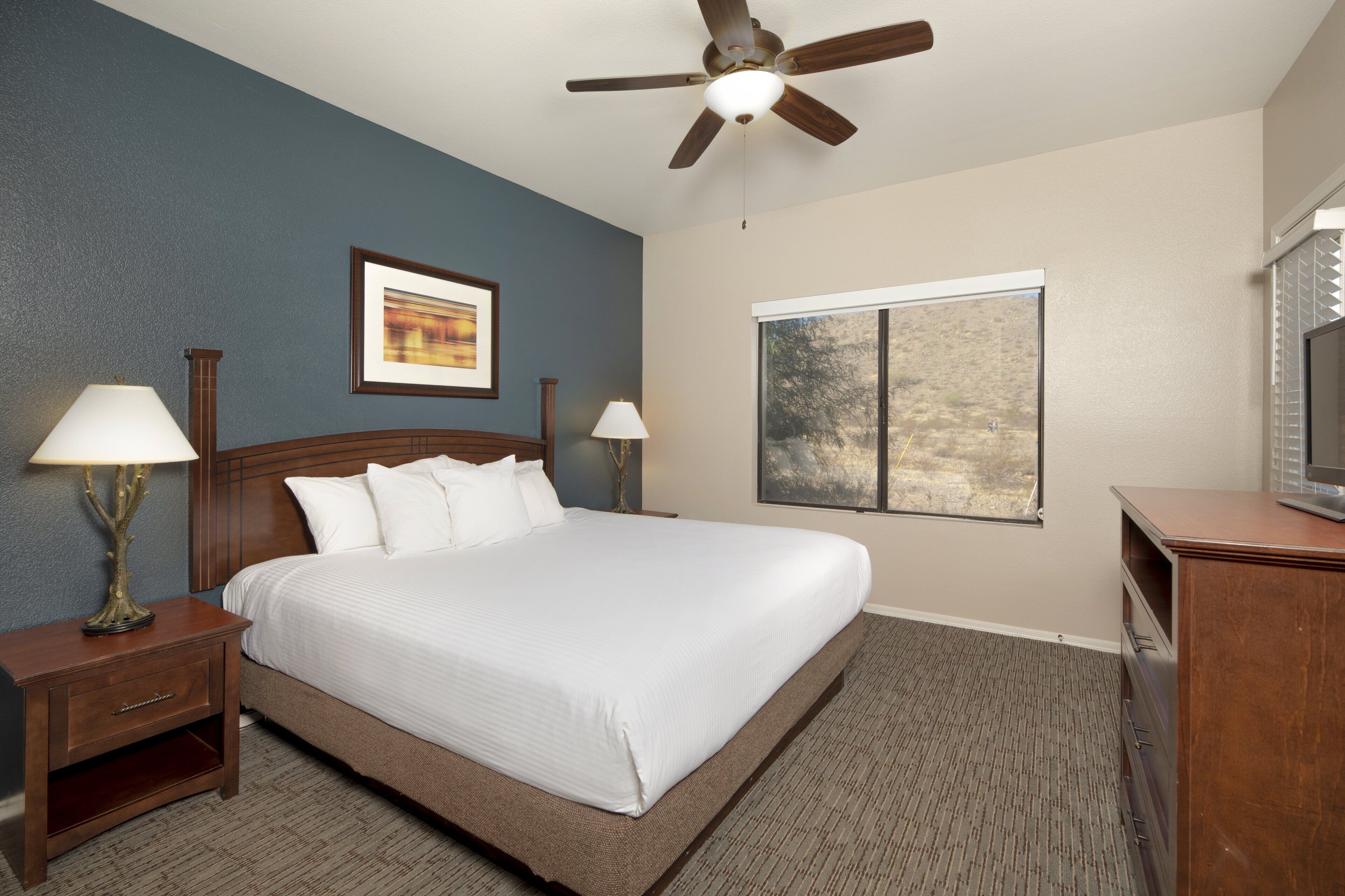 A hotel room with a large bed, two bedside tables with lamps, a ceiling fan, a window with a view, and a TV on a wooden cabinet.