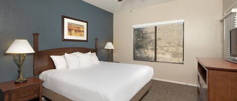 A hotel room with a large bed, two bedside tables with lamps, a ceiling fan, a window with a view, and a TV on a wooden cabinet.