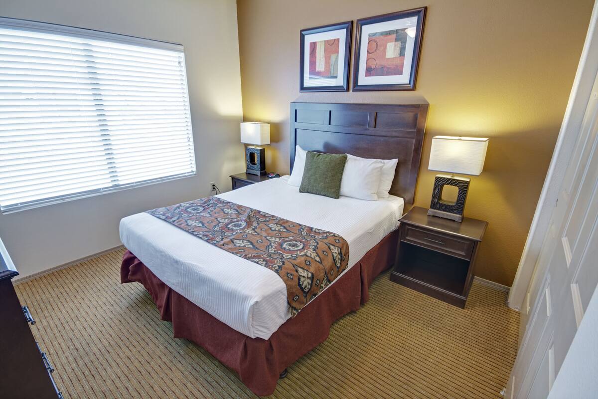 A hotel room with a large bed, two bedside tables with lamps, a window with blinds, and two framed pictures on the wall.