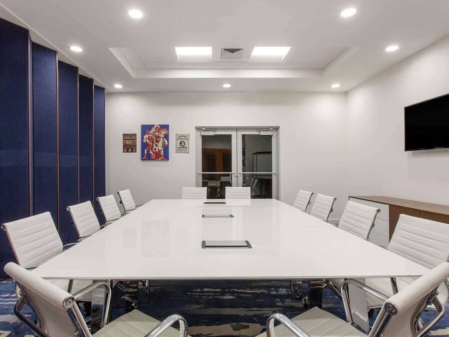 A conference room with a long white table, white chairs, a flat-screen TV, and a door with glass panels.