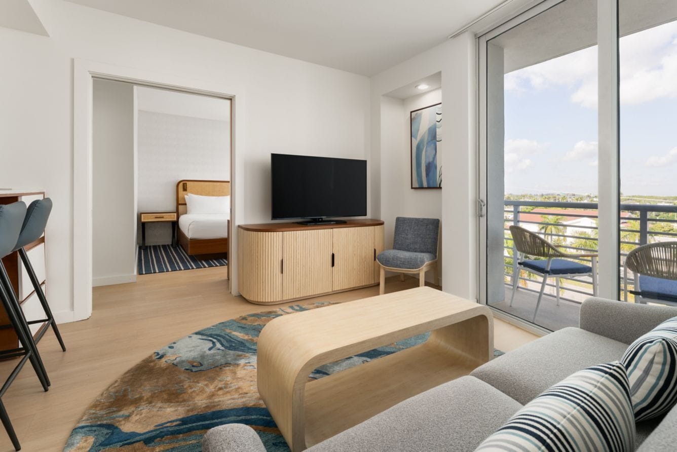 Habitación de hotel moderna con cama, televisión, zona de estar con sofá y sillones, aparador de madera y balcón con mobiliario exterior.