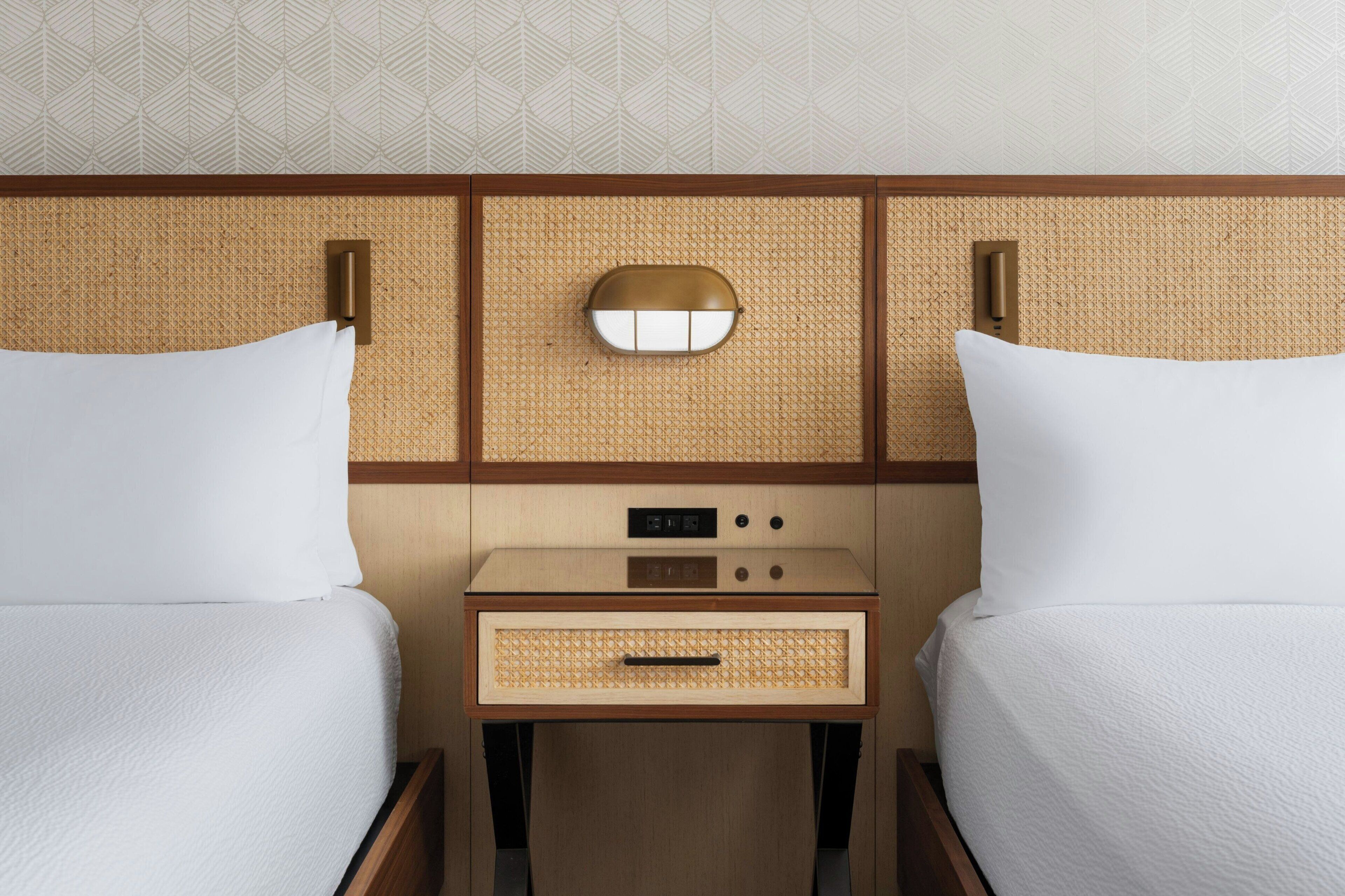 A hotel room with two beds, a bedside table with a lamp, and a wall-mounted light fixture.