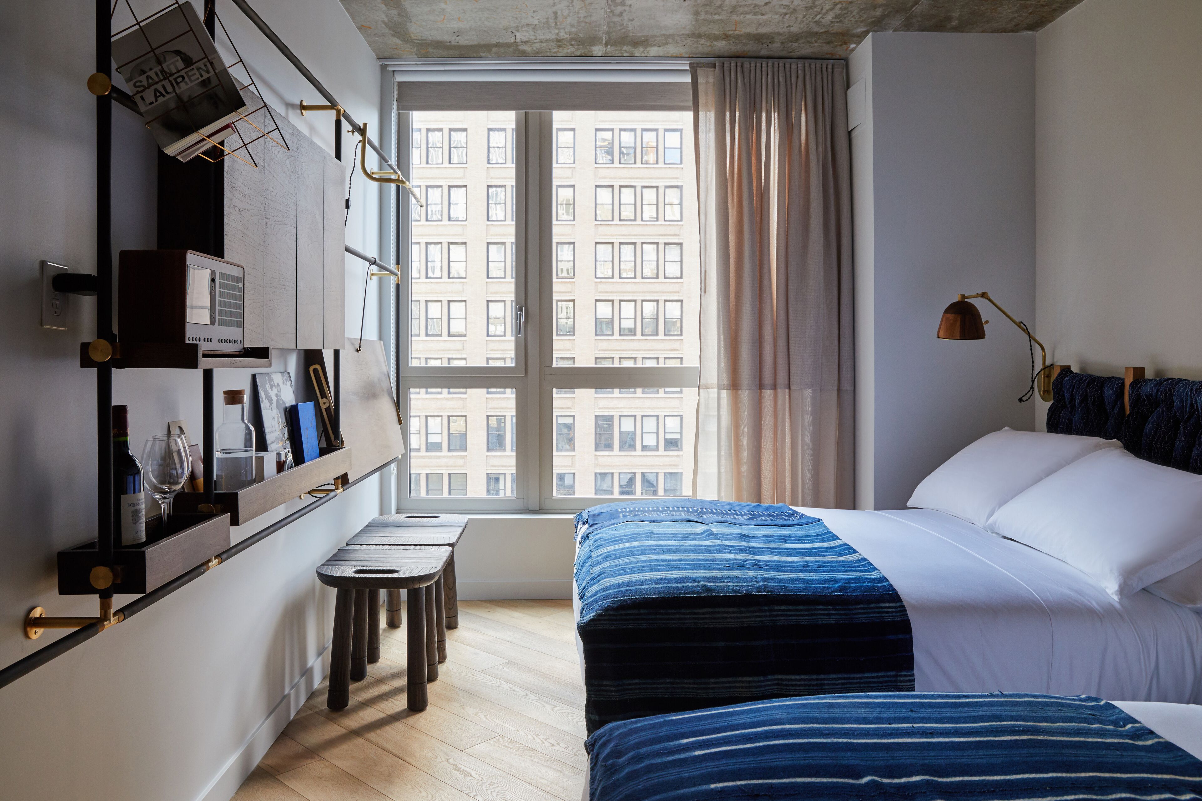 A modern hotel room with a large window, a bed with blue bedding, a bedside table, and a small desk with a lamp.