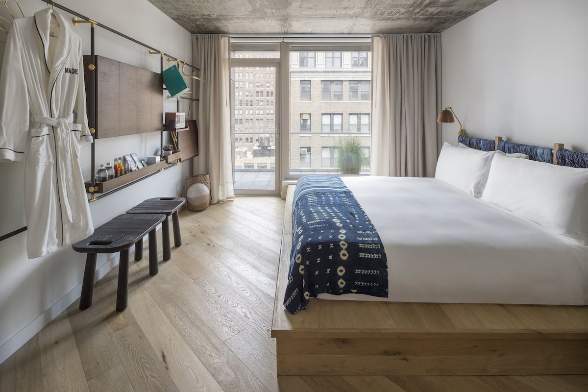 Habitación de hotel con una cama grande, una banca, un perchero y vistas a la ciudad a través de la ventana.