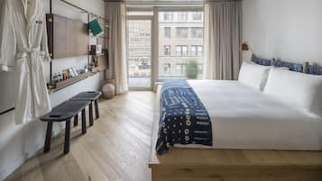 A hotel room with a large bed, a bench, a robe rack, and a view of the city through the window.