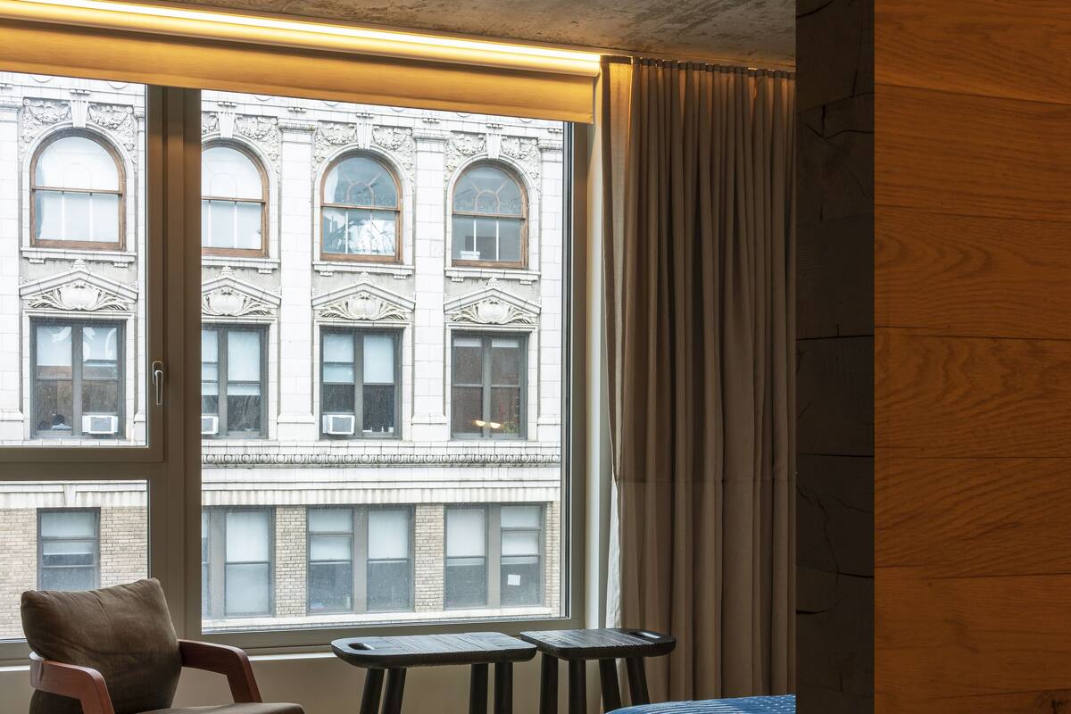 Una habitación con una cama, una silla, una mesita y vistas a un edificio a través de un ventanal grande.