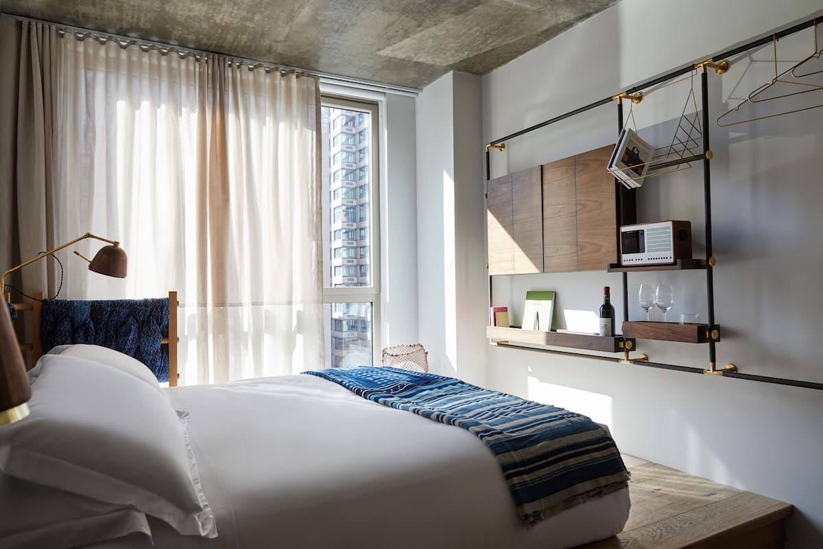 Un dormitorio moderno con una cama grande, una mesita de noche, una lámpara colgante, una ventana con cortinas y un estante de pared con una radio y vasos.