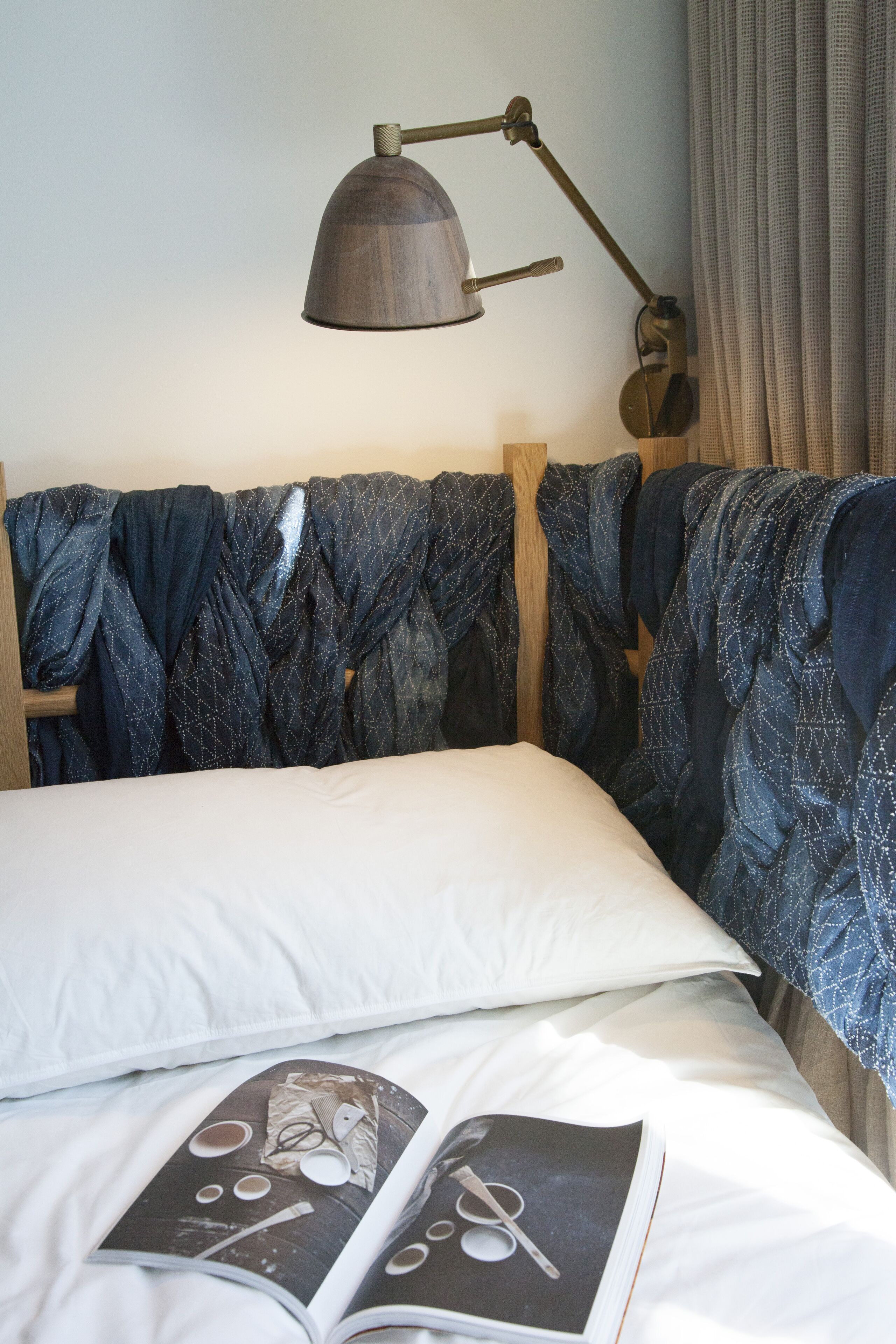 A neatly made bed with a white pillow and a magazine on it. There is a wooden headboard with a blue patterned blanket draped over it. A brown floor lamp is positioned above the bed.