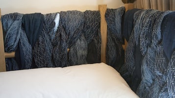 A neatly made bed with a white pillow and a magazine on it. There is a wooden headboard with a blue patterned blanket draped over it. A brown floor lamp is positioned above the bed.