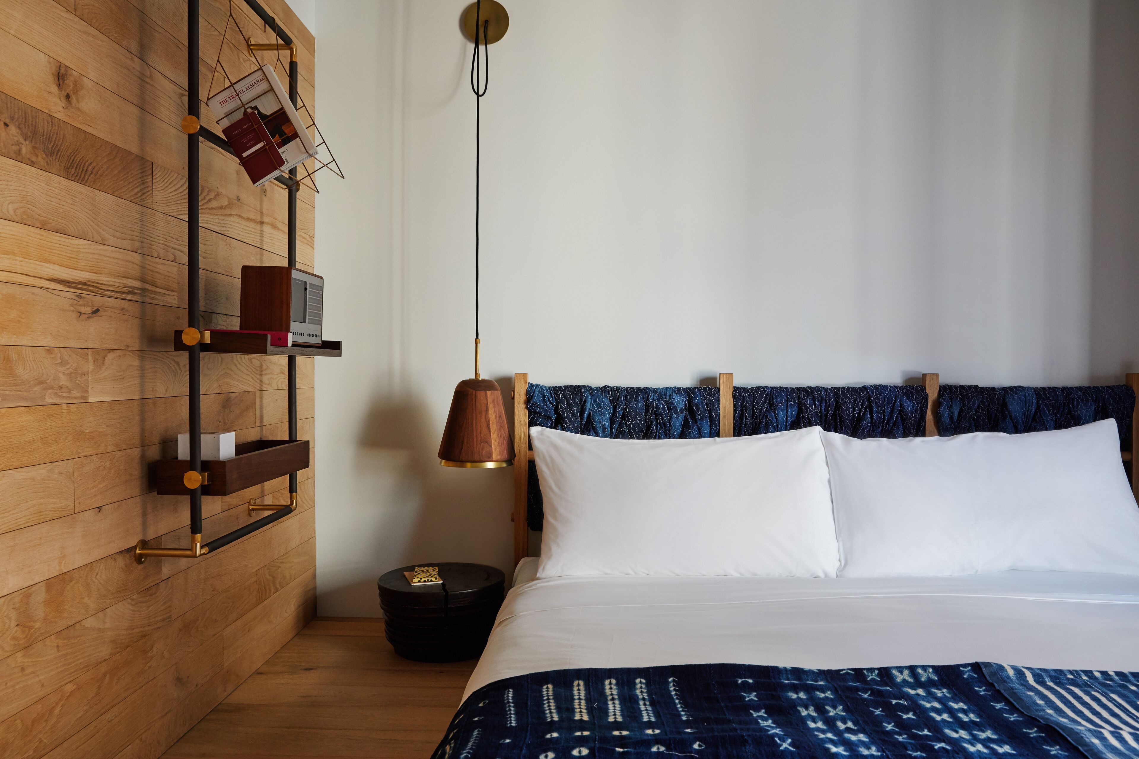 A hotel room with a wooden wall, a bed with white linens, a blue patterned blanket, a wooden nightstand with a lamp, and a shelf with books and a remote control.