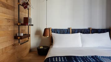 A hotel room with a wooden wall, a bed with white linens, a blue patterned blanket, a wooden nightstand with a lamp, and a shelf with books and a remote control.