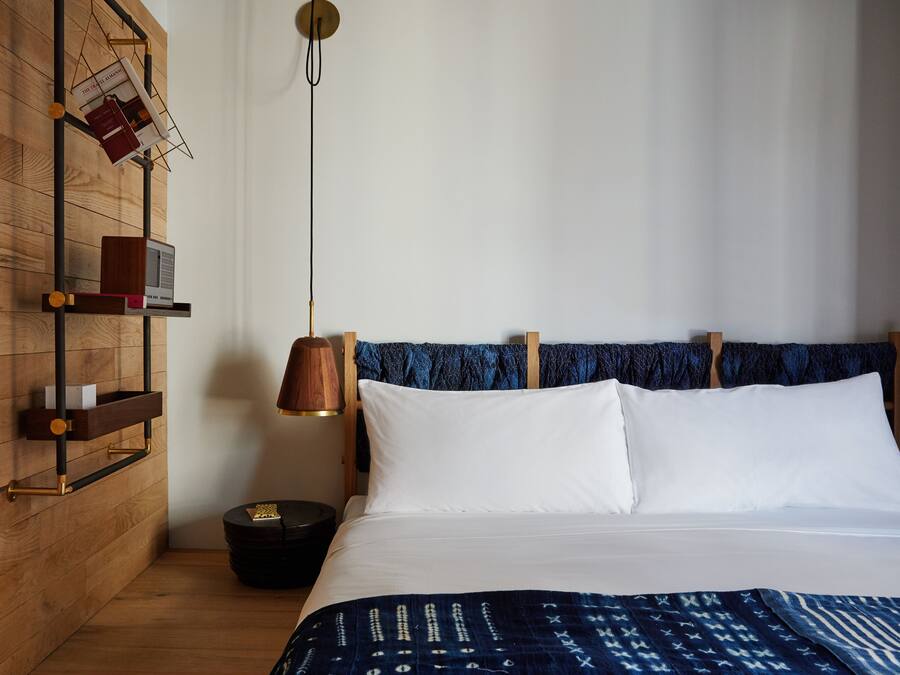 A hotel room with a wooden wall, a bed with white linens, a blue patterned blanket, a wooden nightstand with a lamp, and a shelf with books and a remote control.