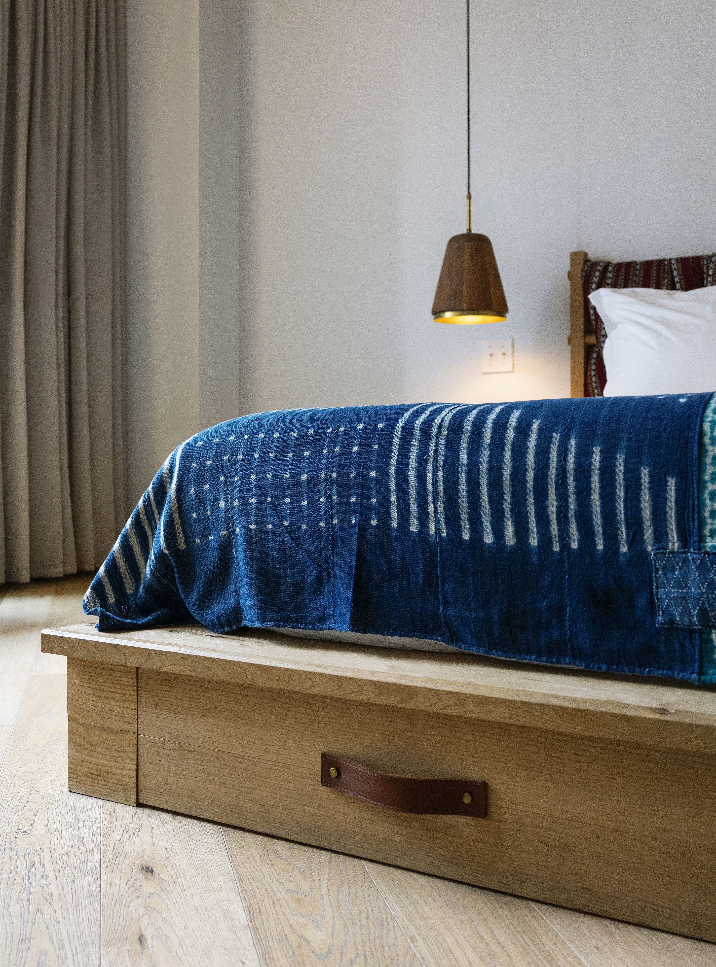 Una cama con un edredón azul, cabecera de madera, una lámpara colgante y una mesita de noche de madera con un cajón.