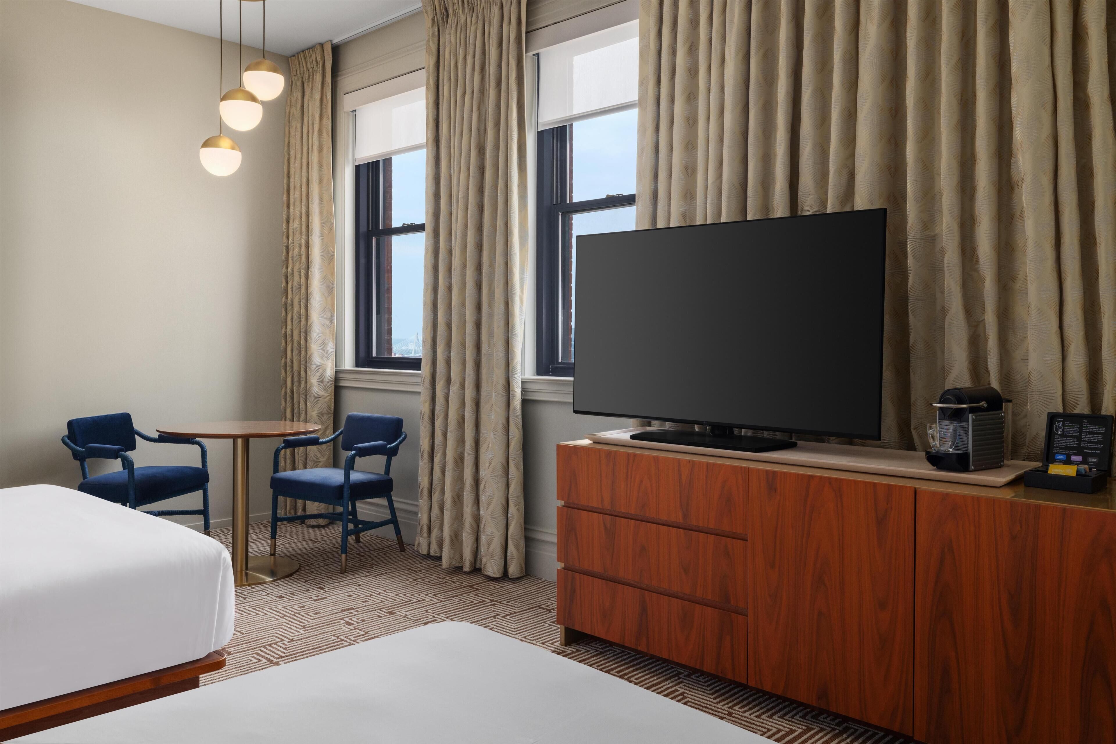 A hotel room with a bed, a television on a wooden cabinet, two chairs, a small table, and a window with curtains.