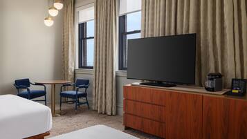 A hotel room with a bed, a television on a wooden cabinet, two chairs, a small table, and a window with curtains.