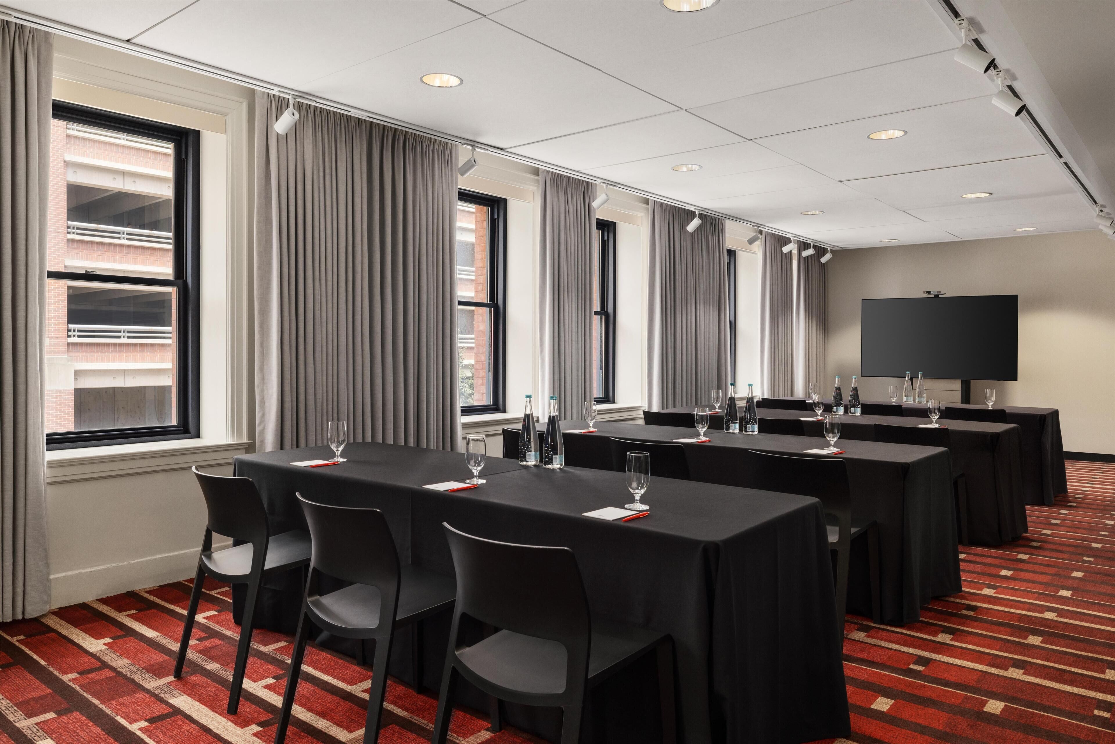 A conference room with multiple tables and chairs, a large screen, and windows with curtains.