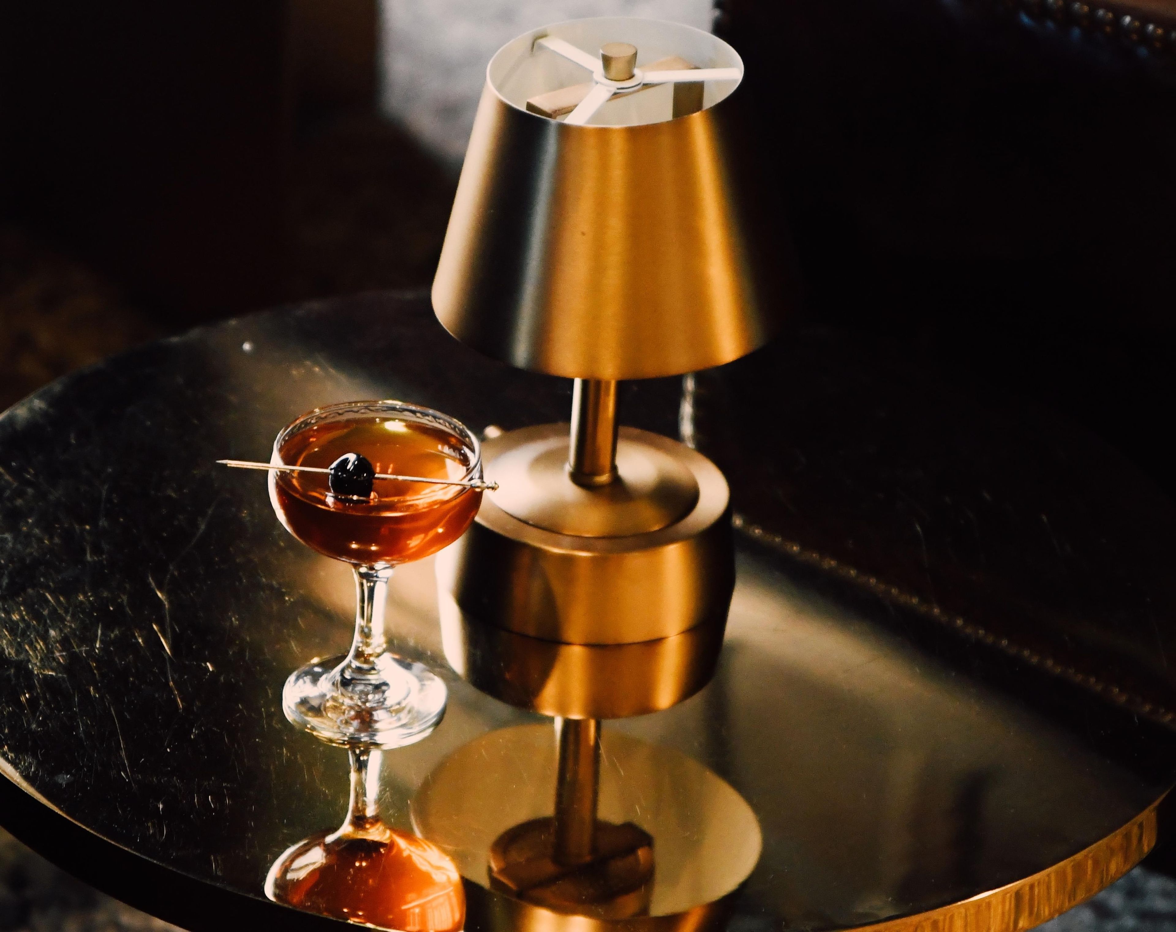 A cocktail glass with a garnish on a reflective surface, a metallic lamp, and a blurred background.