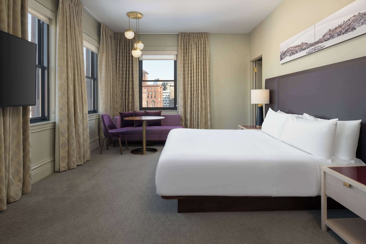 Une chambre d’hôtel avec un grand lit, une télévision, un canapé, une petite table et une vue sur les bâtiments par la fenêtre.