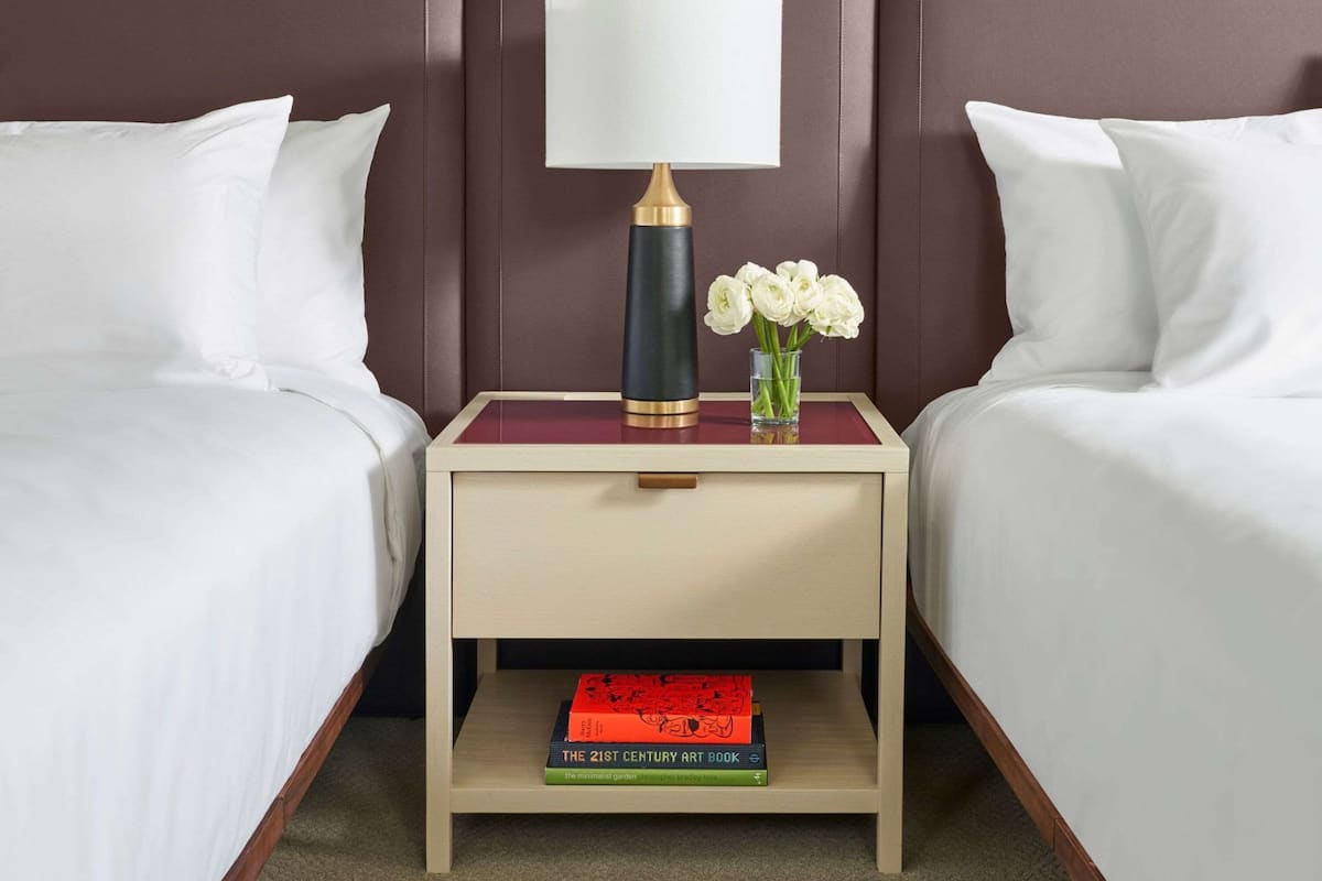 Une chambre d’hôtel avec deux lits, une table de chevet avec une lampe, un vase avec des fleurs et un livre sur la table de chevet.