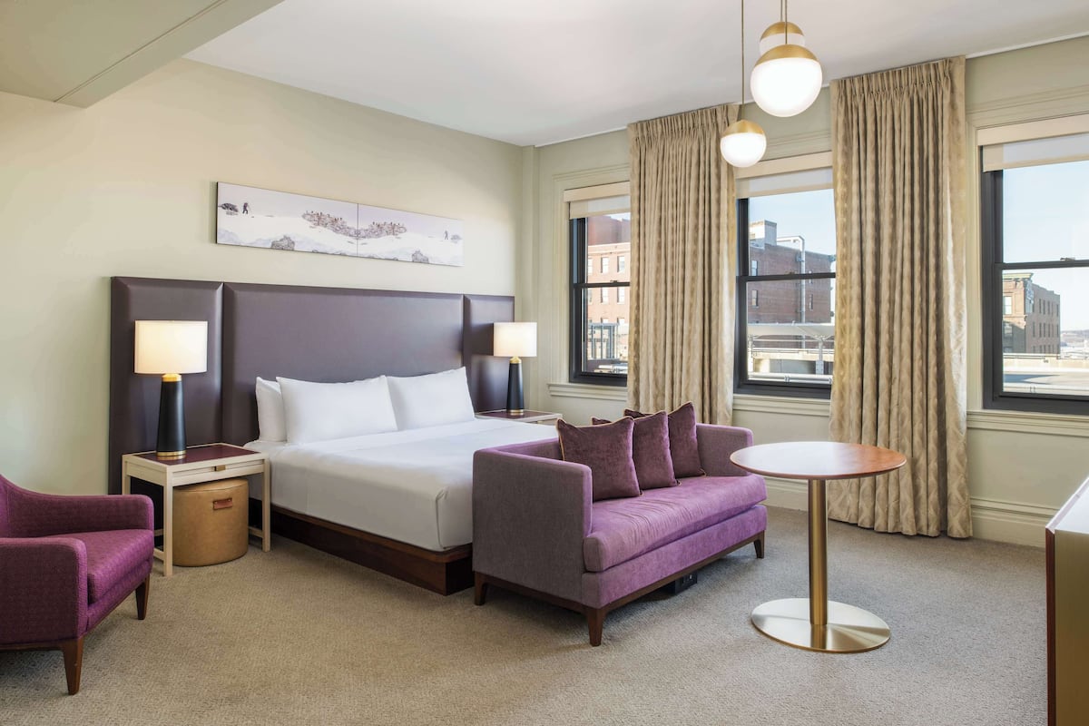 Une chambre d’hôtel avec un grand lit, un fauteuil violet, une table ronde et une vue sur la ville grâce à de grandes fenêtres.
