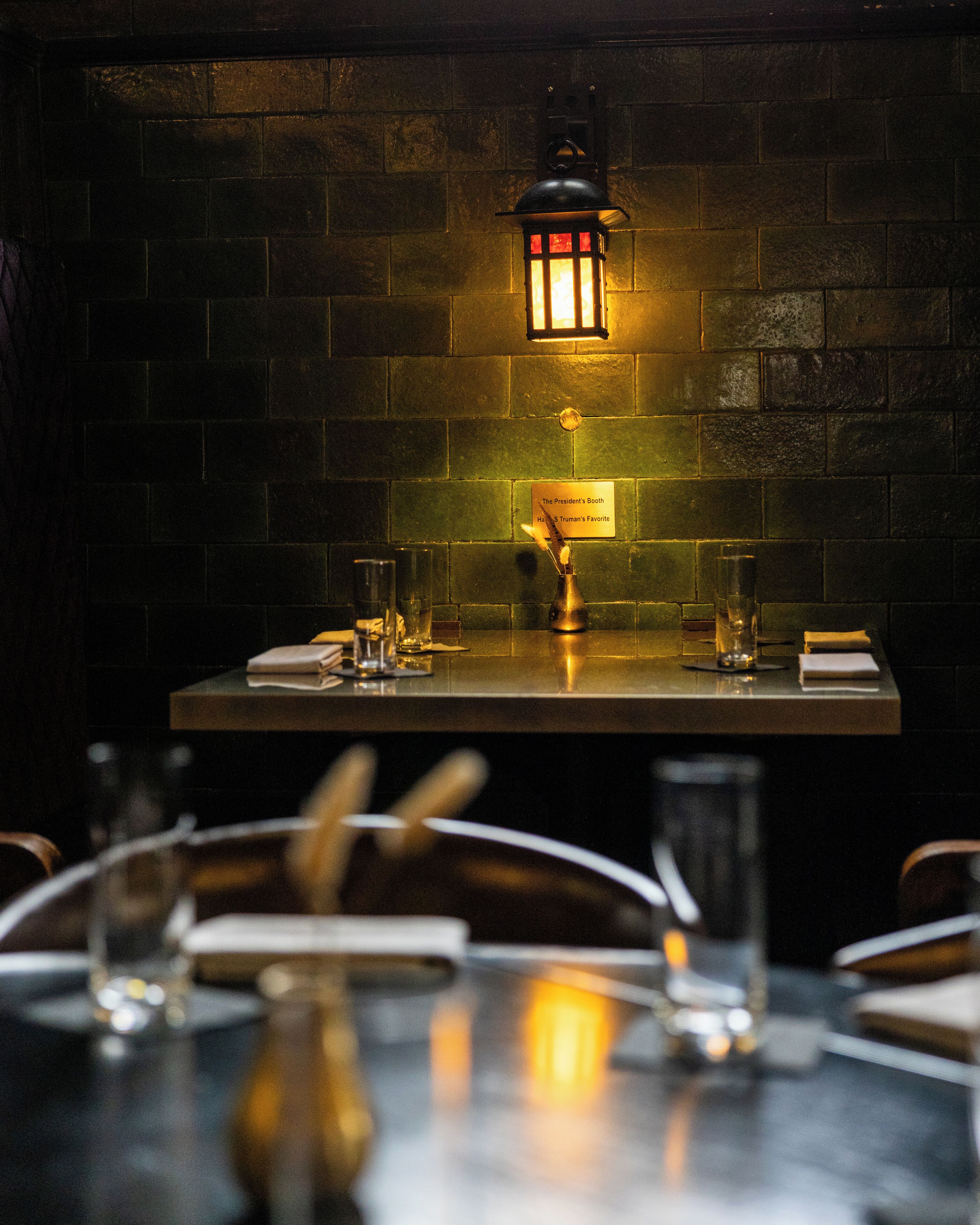 A dimly lit restaurant interior with a brick wall, a table set with glasses and cutlery, and a sign indicating a booth.
