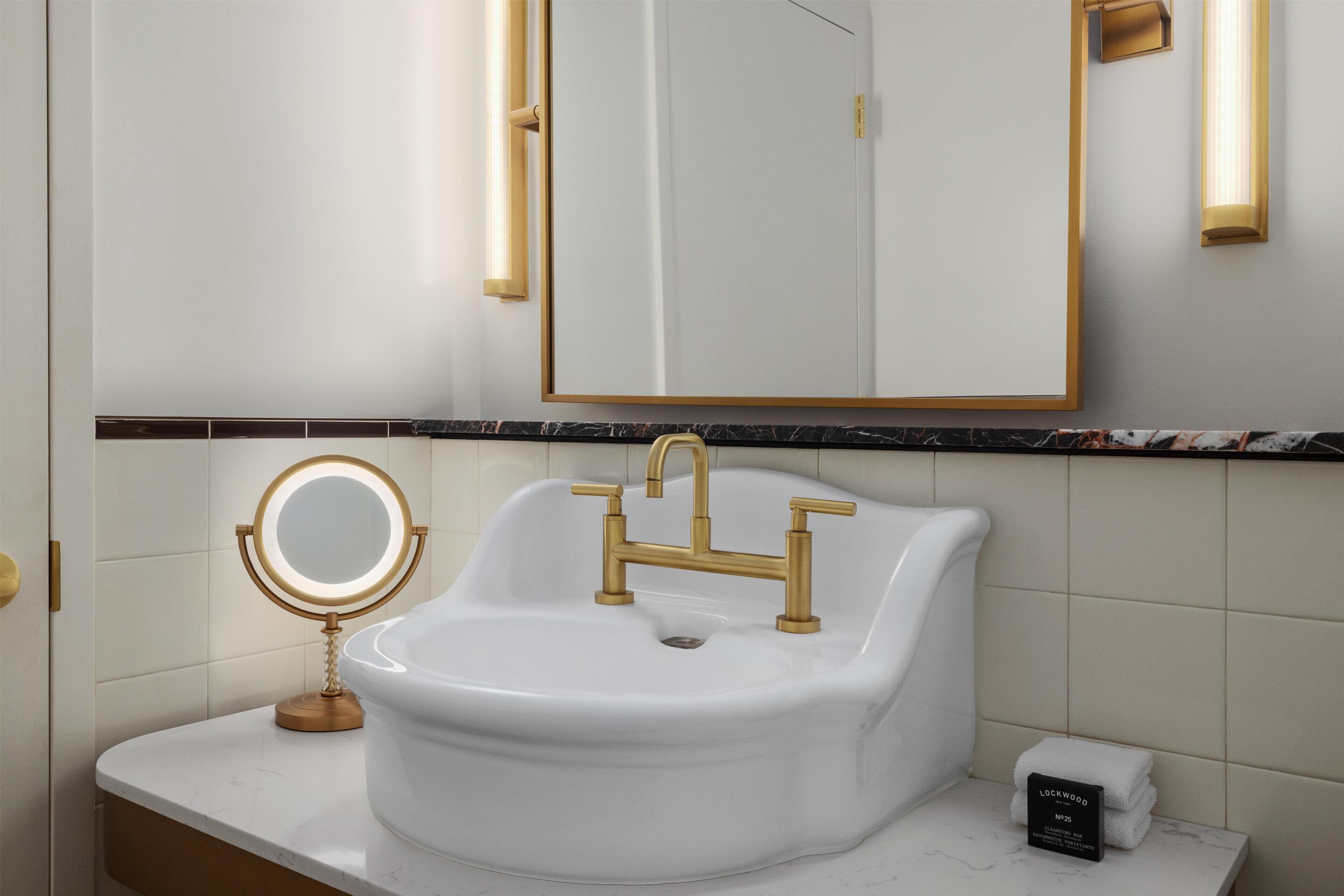 A bathroom with a white sink, gold faucets, and a round mirror with a light.