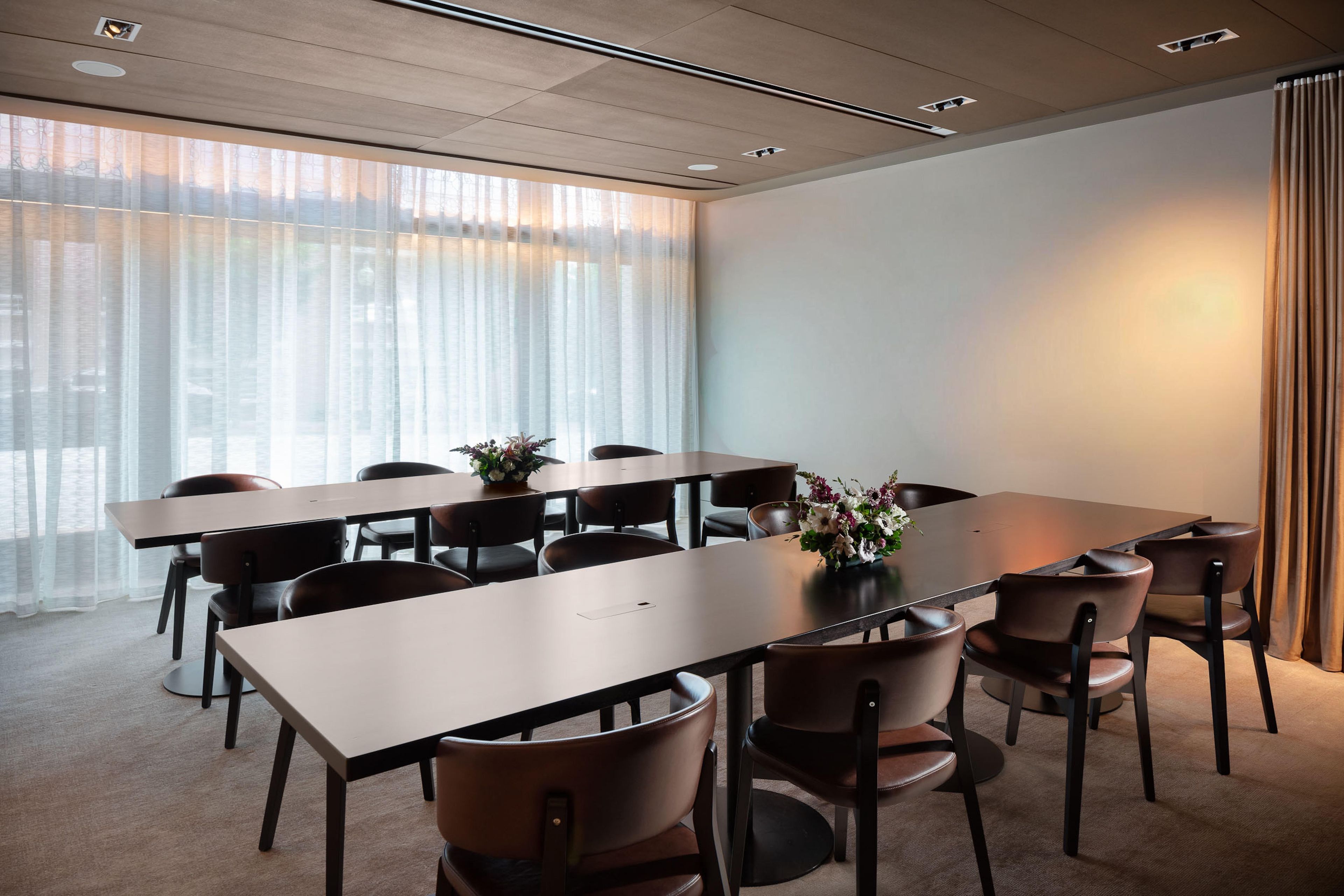 A conference room with a long table and chairs, large windows with sheer curtains, and a carpeted floor.