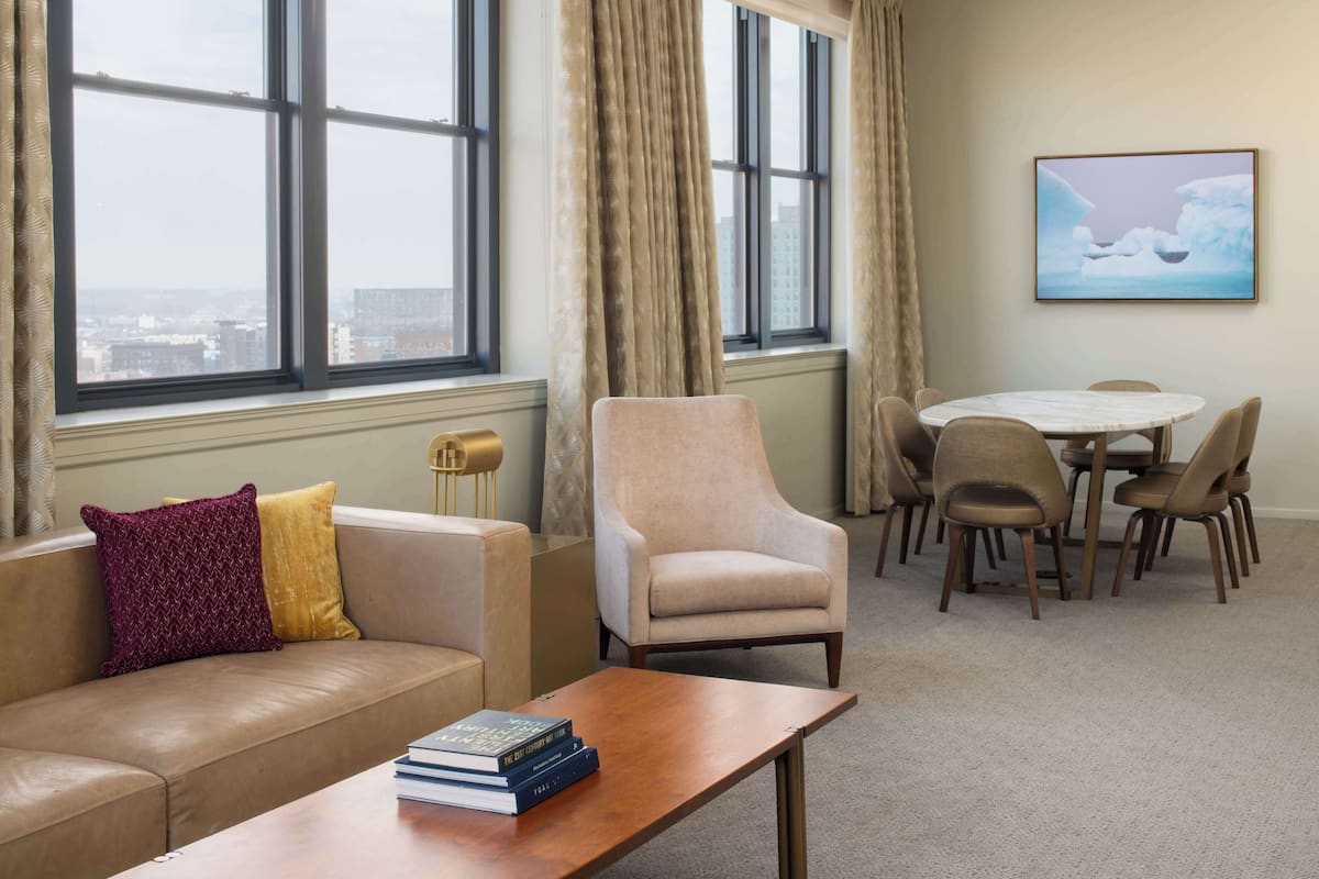 Une chambre d’hôtel avec un canapé en cuir, une table basse en bois sur laquelle sont posés des livres, un fauteuil beige, une table à manger ronde avec des chaises, et une grande fenêtre avec des rideaux.