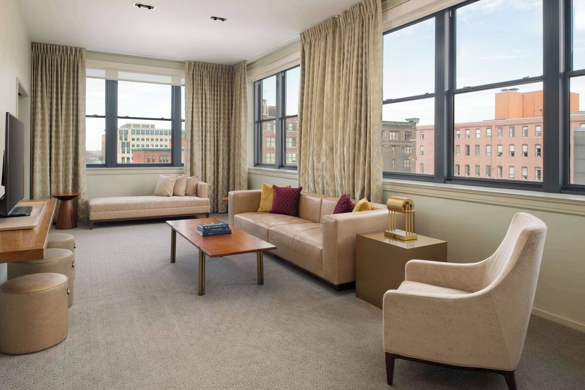 Une chambre d’hôtel moderne, dotée d’un canapé, d’un fauteuil et d’une petite table. De grandes fenêtres offrent une vue sur le paysage urbain.
