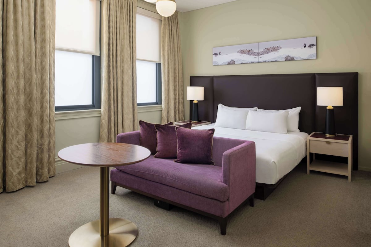 Une chambre d’hôtel avec un lit, deux tables de chevet avec des lampes, un canapé violet, une table ronde et une fenêtre avec des rideaux.