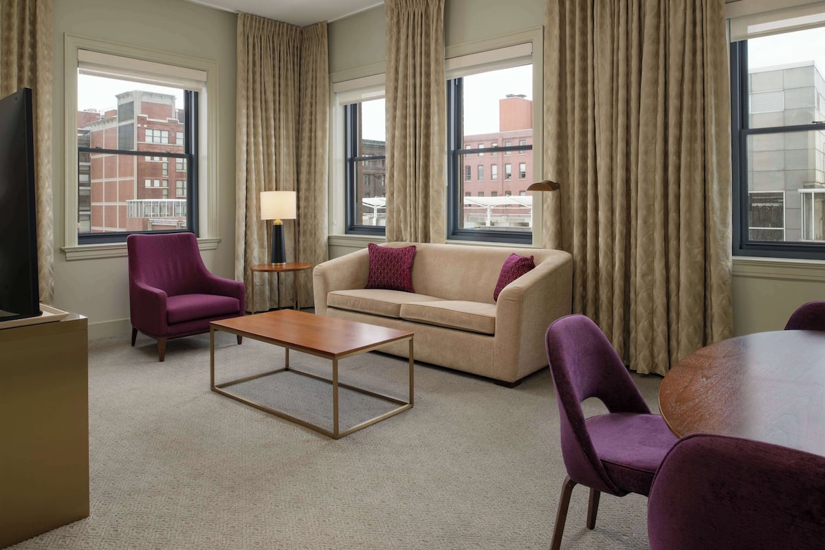 Une chambre d’hôtel avec un canapé beige, un fauteuil violet, une table basse en bois et une table ronde avec des chaises violettes.