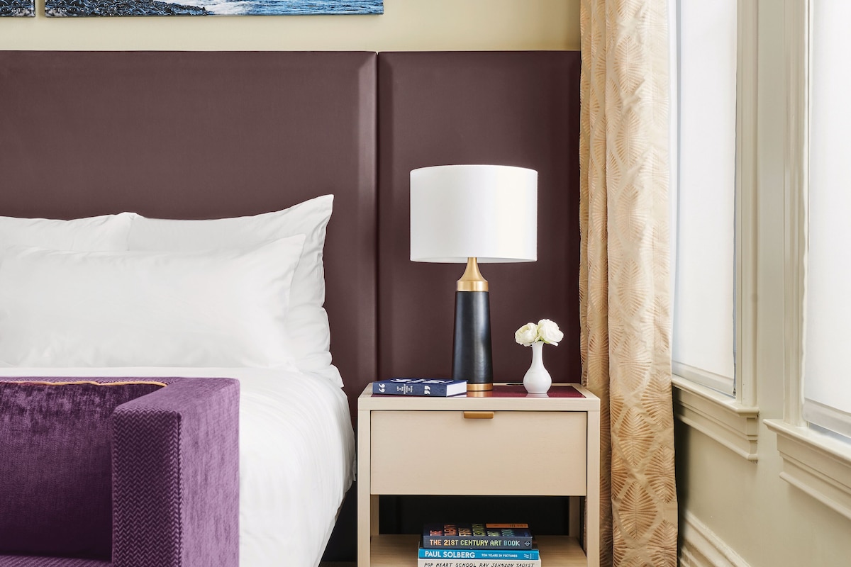 Una habitación de hotel con una cama, un sillón morado, una mesita de noche con una lámpara, libros y un cuadro de una playa.