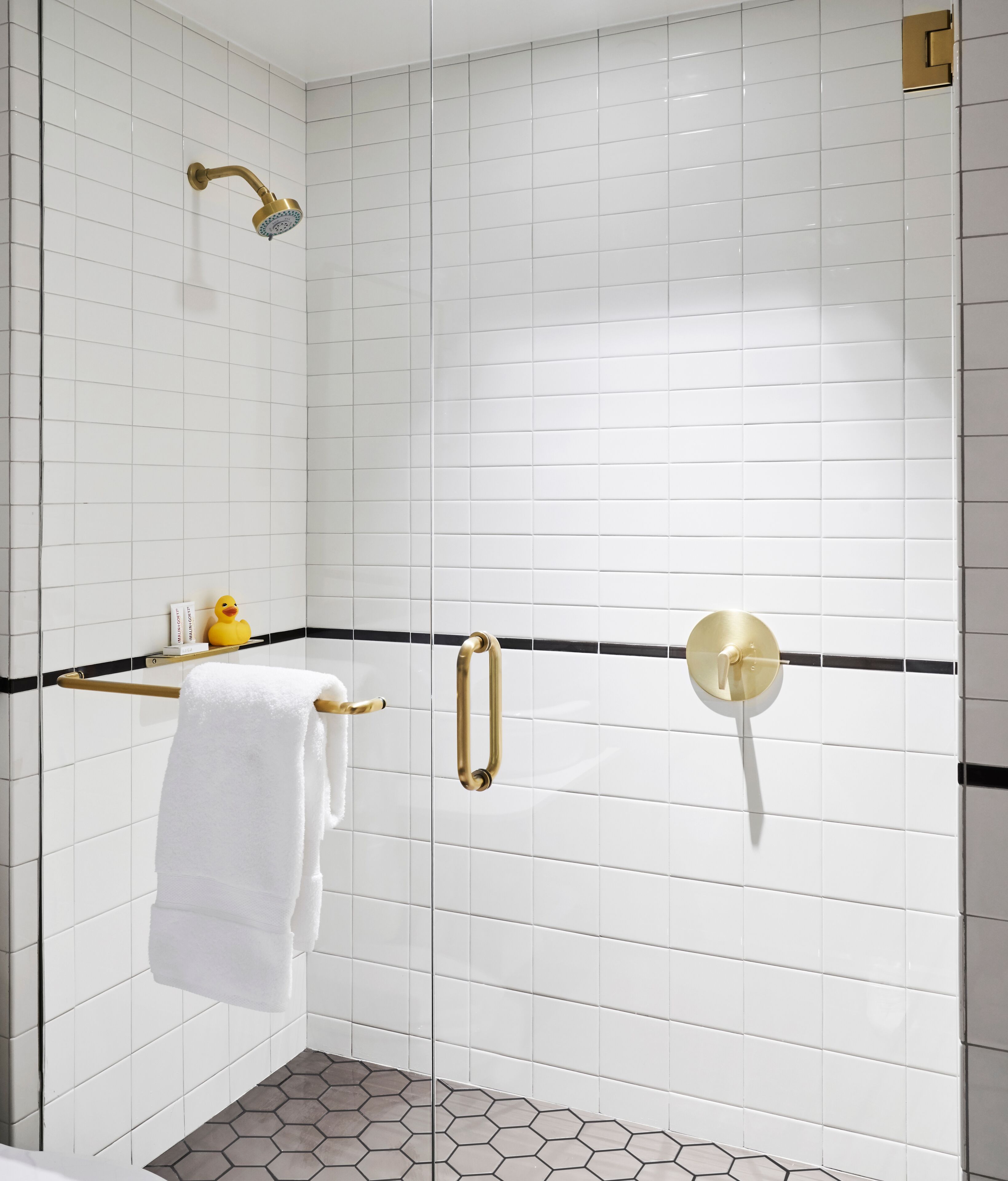 A modern bathroom with white subway tiles, a black and gold shower, a white towel on a wooden rack, and a yellow rubber duck.