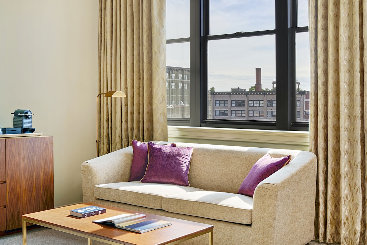 Una sala de estar con un sofá beige, almohadas moradas, una mesa de centro de madera y vistas a edificios a través de un ventanal con cortinas beige.