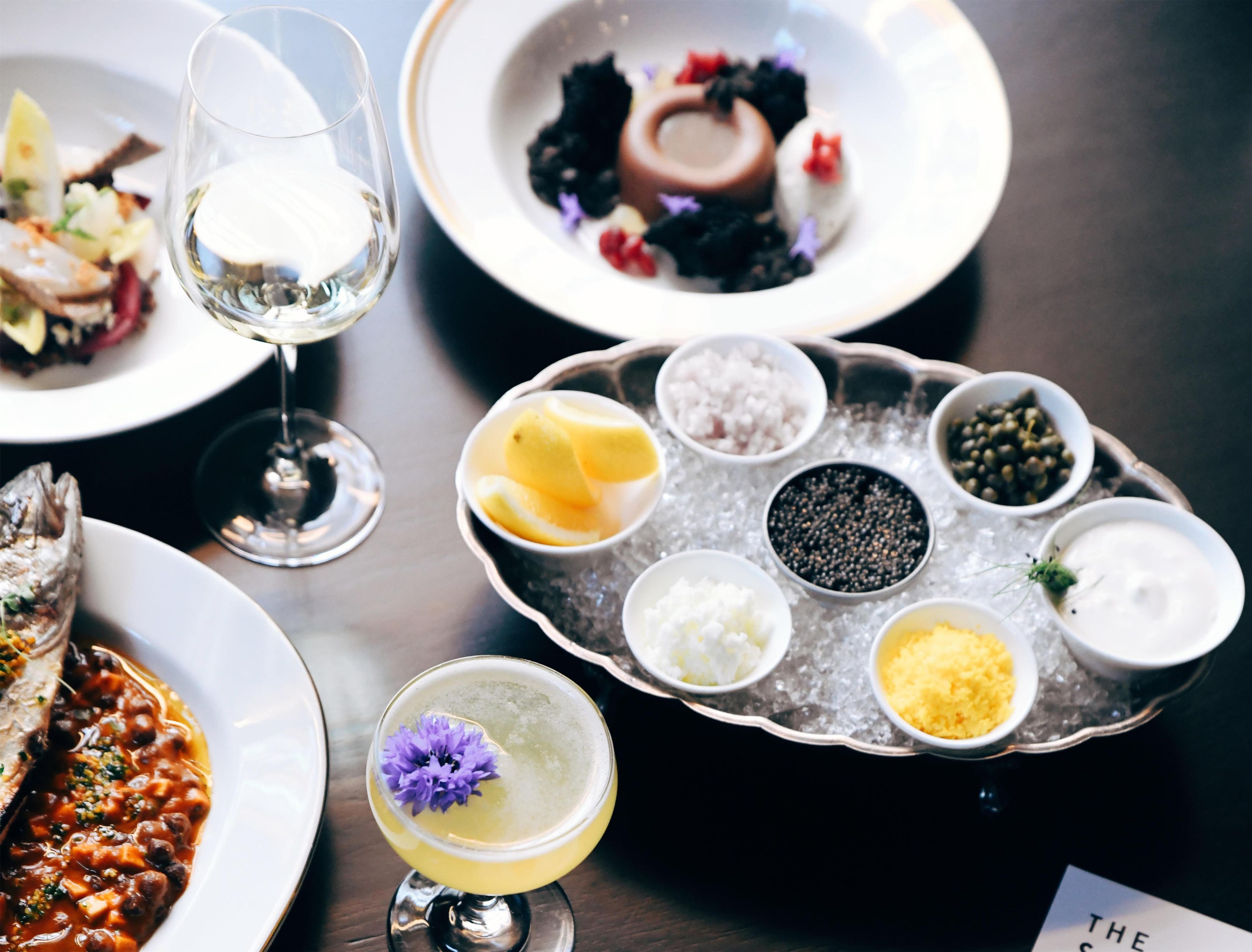 A dining table with various dishes, a glass of white wine, and a tray with condiments.