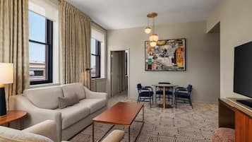 Habitación de hotel con sofá, mesa de centro, televisor, zona de comedor con sillas y alfombra estampada.