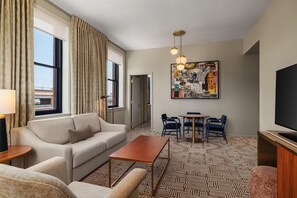 Habitación de hotel con sofá, mesa de centro, televisor, zona de comedor con sillas y alfombra estampada.