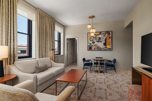 A hotel room with a sofa, a coffee table, a TV, a dining area with chairs, and a patterned rug.