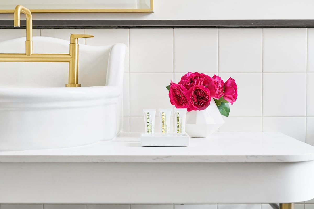 Un baño con lavamanos blanco, detalles dorados y un florero con flores rosadas en la encimera.