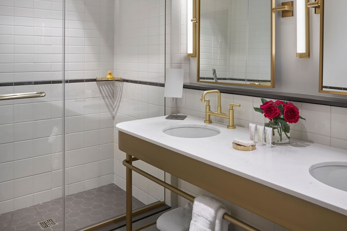 Un baño moderno con ducha envuelta en vidrio, dos lavamanos con detalles dorados y un espejo grande con moldura dorada.
