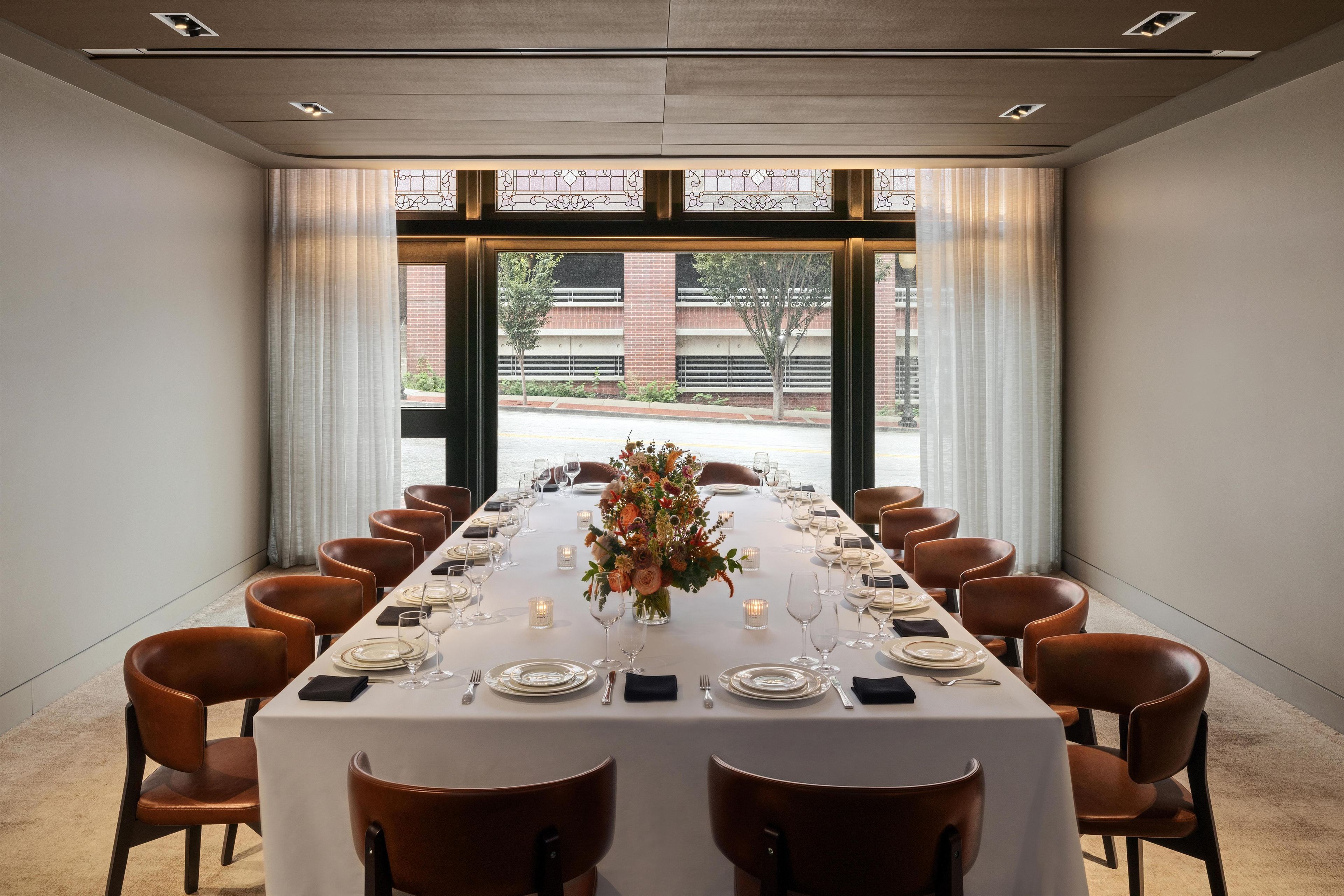 Una larga mesa de comedor dispuesta para una comida, con sillas a ambos lados, ventanas grandes con cortinas y vistas a un edificio.
