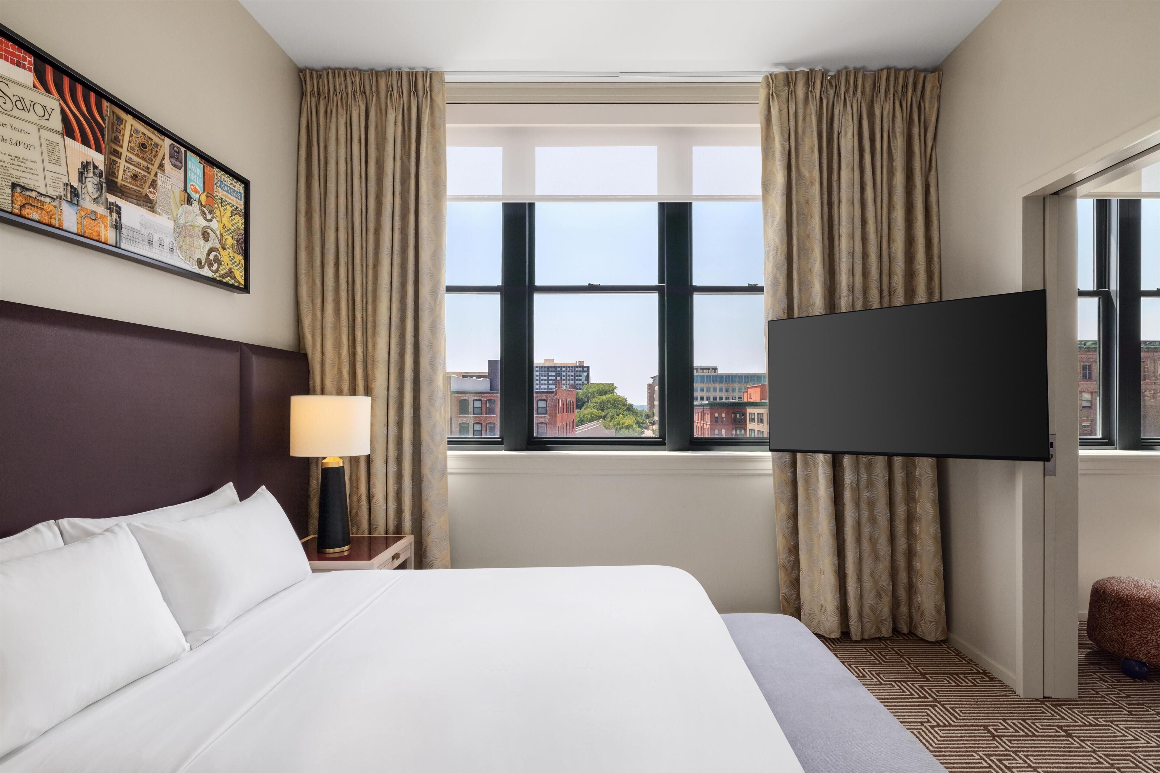 A hotel room with a large bed, a flat-screen TV mounted on the wall, and a window with curtains.