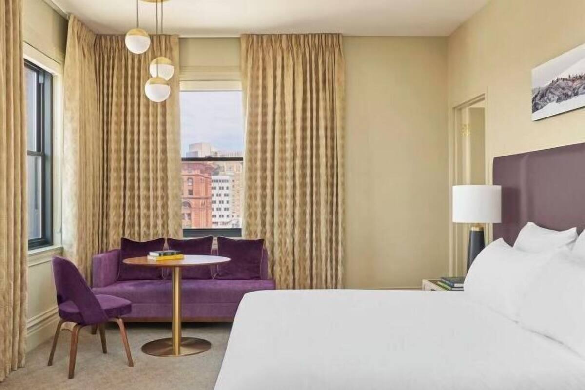 Une chambre d’hôtel avec un lit, un canapé violet, une table ronde et une vue sur les bâtiments environnants.