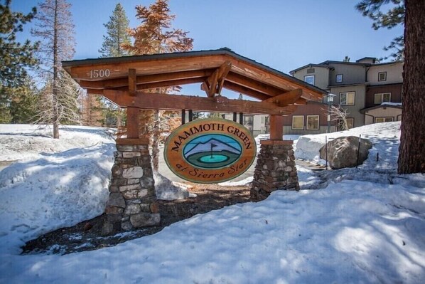 A wooden pergola with a stone base, a circular sign with the text "Mammoth Green at Sierra Star", and a snow-covered landscape with trees and buildings in the background.
