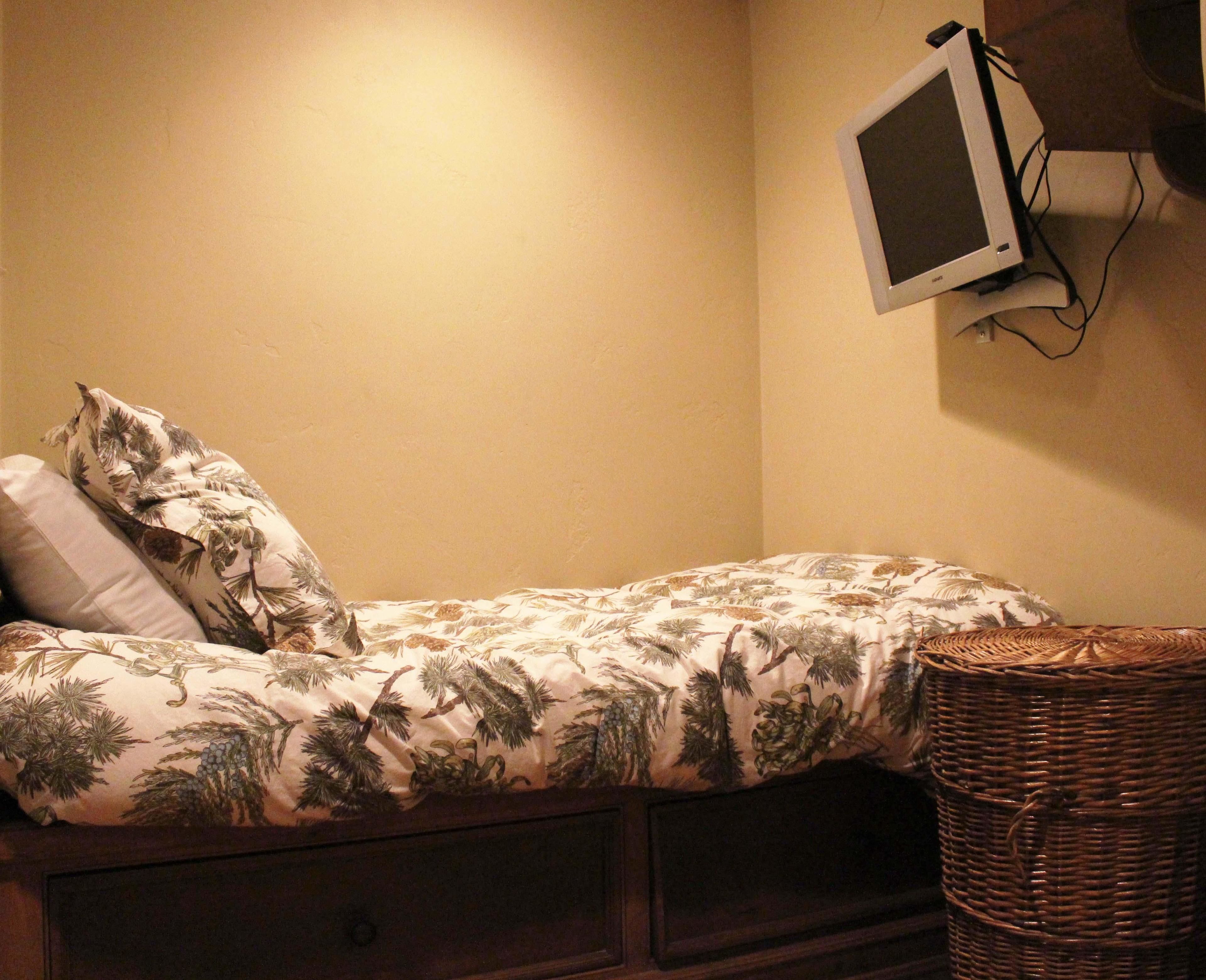 Ein kleines Zimmer mit einem Einzelbett, einer gestreiften Bettdecke, einem Korbwäscheschrank und einem an der Wand befestigten Fernseher.