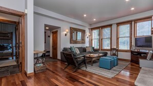 A spacious living room with a sofa, coffee table, and ottoman, wooden flooring, and large windows with blinds. - Snowcreek 883 Phase V (Mammoth Lakes)